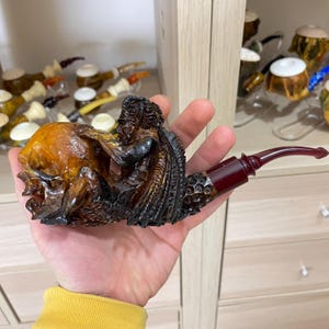 Puede incluir: Una pipa de tabaco tallada con una figura detallada con una capa marrón oscuro y un diseño de calavera. La pipa tiene un tallo marrón rojizo y una boquilla curva. Otras pipas son visibles en el fondo.