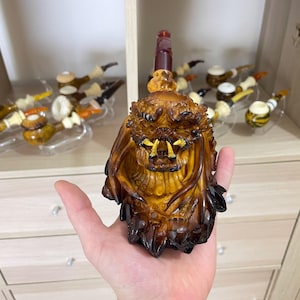 Könnte beinhalten: Eine handgeschnitzte Tabakpfeife mit Predator-Design. Die Pfeife ist überwiegend braun mit gelben Zähnen und einem dunkelroten Mundstück. Die detaillierte Schnitzerei zeigt das Gesicht und den Kopf des Predators. Ca. 15 cm lang.