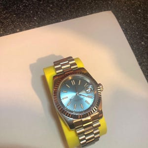 Puede incluir: Un reloj de pulsera de acero inoxidable con esfera azul claro y bisel estriado. El reloj tiene una correa plateada y se muestra sobre un soporte de reloj amarillo. La esfera del reloj presenta marcadores de hora y agujas plateadas.