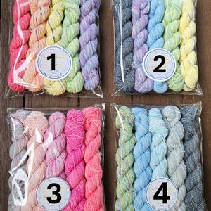 Könnte beinhalten: Vier Packungen bunter Garnknäuel in durchsichtigen Plastiktüten. Jede Packung hat eine andere Farbpalette, darunter Regenbogenfarben, Blau-, Rosa- und Grüntöne. Jede Packung hat einen runden Aufkleber mit einer Nummer.