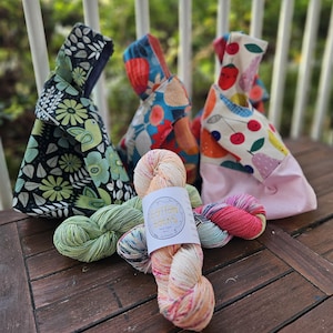 Op de afbeelding: Drie stoffen knooptassen met diverse patronen, waaronder zwart-groene bloemen, blauwe en oranje abstracte vormen, en wit met rode kersen en peren, naast een streng oranje, roze en groen gemêleerd garen met een Cotton Socks label.