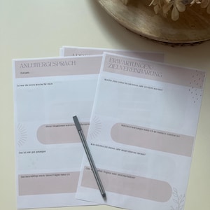 Op de afbeelding: Een stapel bedrukte vellen met Duitse tekst, met roze en witte ontwerpen. Een zilveren pen ligt op de pagina's. De vellen zijn bedoeld voor doelstellingen en zelfreflectie, met prompts voor journaling.