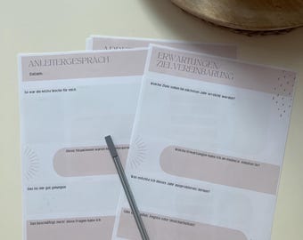 May include: A stack of printed worksheets with German text, featuring pink and white designs. A silver pen rests on the pages. The worksheets are for goal setting and self-reflection, with prompts for journaling.