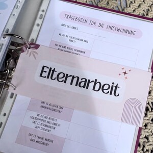 Könnte beinhalten: Ein rosa-weißer Ordner mit einem Reiter mit der Aufschrift "Elternarbeit". Der Ordner enthält einen Fragebogen mit dem Titel "Fragebogen für die Eingewöhnung" mit Fragen zur Lebenssituation des Kindes und seinen Erfahrungen in der Kita.