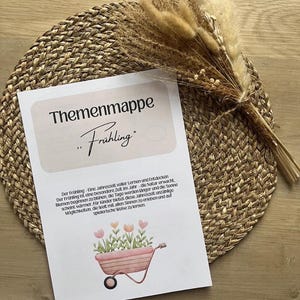 Puede incluir: Una tarjeta impresa con el texto alemán "Themenmappe Frühling", con una ilustración de acuarela de una carretilla llena de flores. La tarjeta descansa sobre un mantel individual tejido, con hierbas y flores secas en el fondo.
