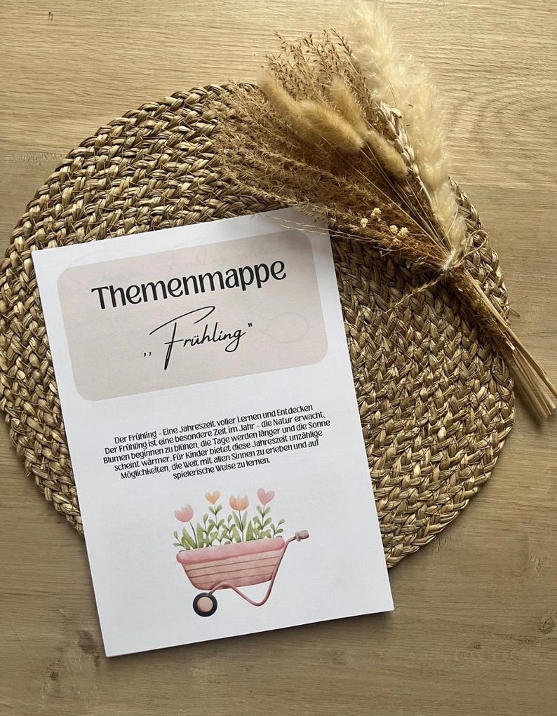 May include: A printed card with the German text "Themenmappe Fr&uuml;hling" featuring a watercolor illustration of a wheelbarrow filled with flowers. The card rests on a woven placemat, with dried grasses and flowers in the background.