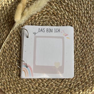 Peut inclure: Un album photo carré relié par deux anneaux métalliques. La couverture présente le texte "DAS BIN ICH" au-dessus d'un cadre rectangulaire marron clair avec une petite icône de cœur, un motif arc-en-ciel et des confettis colorés.