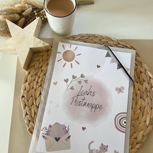 May include: A clear plastic document holder with a decorative insert featuring a sun, hearts, and a cat. The text "Leahs Postmappe" is visible. A cup of coffee, a wooden star, and dried flowers are also present.