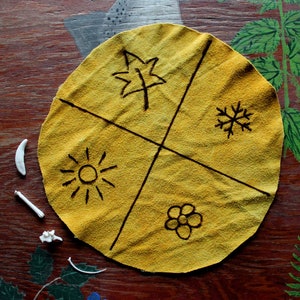 May include: A yellow fabric circle divided into four sections with black lines. Each section has a different symbol drawn in black: a leaf, a snowflake, a sun, and a flower.