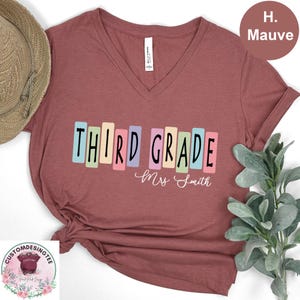 May include: Mauve V-neck t-shirt with the words "THIRD GRADE" in colorful block letters. Below the text is the name "Mrs. Smith" in cursive. The shirt is a soft, comfortable material.