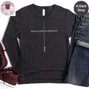 May include: A dark grey long-sleeve shirt featuring a white vertical cross graphic. The text reads THE WAY, THE TRUTH, AND THE LIFE with JOHN 14:6 printed vertically below the cross. The shirt is paired with blue denim jeans and maroon sneakers.