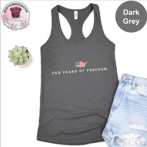 a grey tank top with the text "250 YEARS OF FREEDOM" printed on it, along with an American flag design. The tank top is placed next to a pair of denim shorts, and there is a small potted plant and a round logo in the background.
