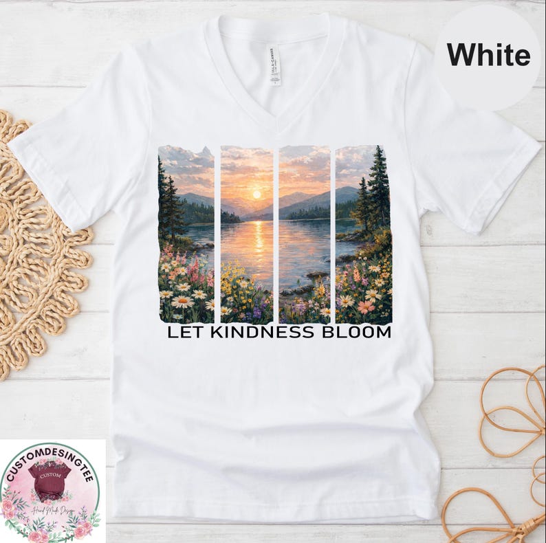 A white t-shirt with a printed design depicting a serene landscape with a lake, mountains, and a sunset. The text "Let Kindness Bloom" is printed below the image.