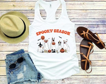 May include: A white racerback tank top featuring the orange text SPOOKY SEASON above a graphic of five dogs dressed as ghosts, surrounded by bats and pumpkins. The tank top is displayed with a straw hat, denim shorts, sunglasses, and sandals.