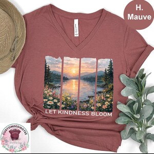 a red v-neck t-shirt with a printed design depicting a serene landscape with a lake, mountains, and a sunset. The text "LET KINDNESS BLOOM" is printed below the image.