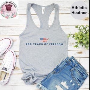 a gray tank top with the text "250 YEARS OF FREEDOM" printed on it, along with a pair of denim shorts and white sneakers. The tank top is displayed on a wooden surface, and there is a plant and a pair of shoes placed next to it.