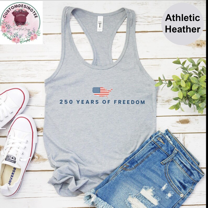 a gray tank top with the text "250 YEARS OF FREEDOM" printed on it, along with a pair of denim shorts and white sneakers. The tank top is displayed on a wooden surface, and there is a plant and a pair of shoes placed next to it.