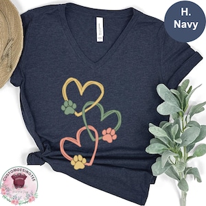 May include: Navy blue V-neck t-shirt featuring a design of three hearts in yellow, green, and coral, each with a paw print. The shirt has a relaxed fit and is made of a soft material. The text "H. Navy" is in a navy blue circle.