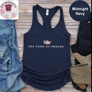 a navy blue tank top with the text "250 YEARS OF FREEDOM" printed on it, along with an American flag design. The tank top is laid out on a wooden surface, surrounded by various clothing items such as jeans, a pair of boots, and a pair of shoes.