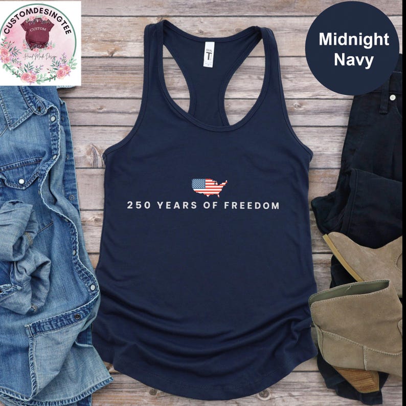 a navy blue tank top with the text "250 YEARS OF FREEDOM" printed on it, along with an American flag design. The tank top is laid out on a wooden surface, surrounded by various clothing items such as jeans, a pair of boots, and a pair of shoes.