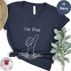 a navy blue t-shirt with a graphic design of a hand giving a thumbs up, accompanied by the text "i'm fine". The t-shirt is placed on a white surface, with a hat and a plant visible in the background.