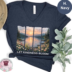 A navy blue v-neck t-shirt with a printed design depicting a serene landscape with a lake, mountains, and a sunset. The text "LET KINDNESS BLOOM" is printed below the image.