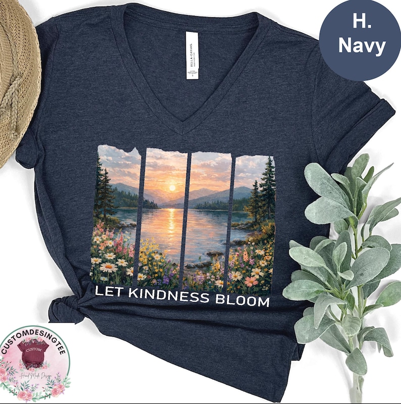 A navy blue v-neck t-shirt with a printed design depicting a serene landscape with a lake, mountains, and a sunset. The text "LET KINDNESS BLOOM" is printed below the image.