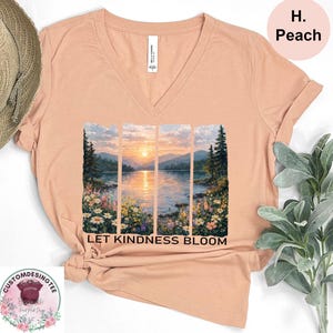 a peach-colored t-shirt with a printed design depicting a serene landscape with a sunset over a body of water. The text "LET KINDNESS BLOOM" is printed below the image.