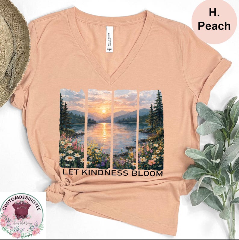 a peach-colored t-shirt with a printed design depicting a serene landscape with a sunset over a body of water. The text "LET KINDNESS BLOOM" is printed below the image.