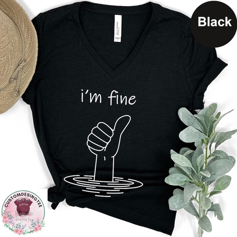 a black t-shirt with a white graphic design depicting a hand giving a thumbs up, surrounded by a water-like shape. The t-shirt is placed on a white surface, with a hat and some greenery visible in the background.