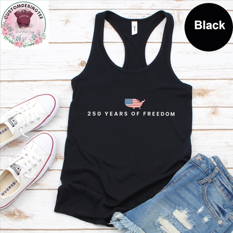 a black tank top with the text "250 YEARS OF FREEDOM" printed on it, along with a graphic of the American flag. The tank top is placed on a wooden surface, and there are a pair of white sneakers and a pair of blue denim shorts visible in the image.