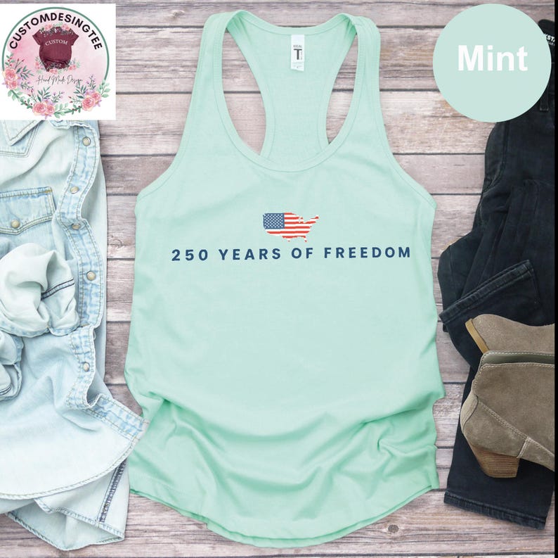 A mint green tank top with the text "250 YEARS OF FREEDOM" printed on it, along with an American flag design, is displayed on a wooden surface alongside a pair of jeans and a pair of boots.