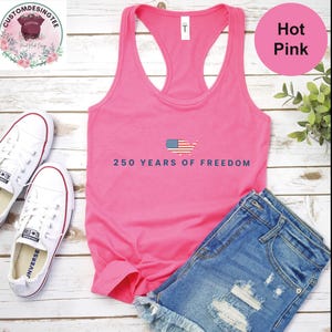A pink tank top with the text "250 YEARS OF FREEDOM" printed on it, along with a pair of denim shorts and white sneakers, all laid out on a wooden surface.