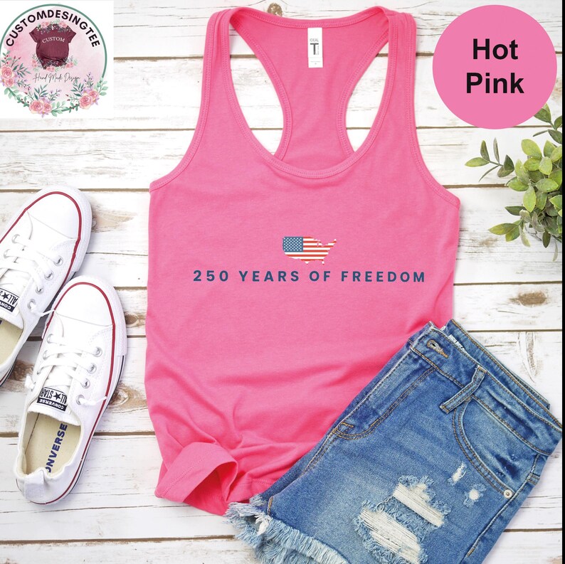 A pink tank top with the text "250 YEARS OF FREEDOM" printed on it, along with a pair of denim shorts and white sneakers, all laid out on a wooden surface.