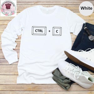 May include: White long-sleeve shirt with a black graphic of a computer keyboard's "CTRL" and "C" keys. The shirt is paired with blue jeans and white sneakers. The shirt is made of a soft, comfortable material.