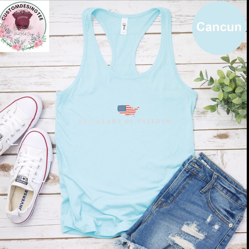 A light blue tank top with the text "250 YEARS OF FREEDOM" printed on it, along with a pair of denim shorts and white sneakers, all laid out on a wooden surface.