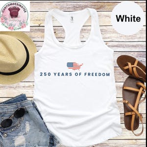 a white tank top with the text "250 YEARS OF FREEDOM" printed on it, along with an American flag design. The tank top is displayed on a wooden surface, accompanied by a straw hat, a pair of sandals, and a pair of jeans.
