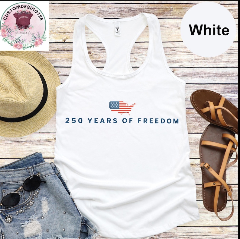 a white tank top with the text "250 YEARS OF FREEDOM" printed on it, along with an American flag design. The tank top is displayed on a wooden surface, accompanied by a straw hat, a pair of sandals, and a pair of jeans.
