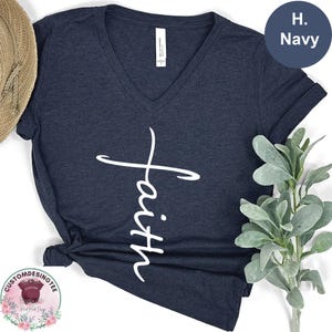 May include: Navy blue V-neck t-shirt with the word "Faith" in a white script. The shirt has short sleeves and a relaxed fit. The shirt is made of a soft material. The color is indicated as "H. Navy".