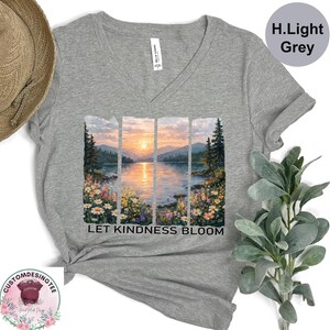 a grey t-shirt with a printed design depicting a serene landscape with a lake, mountains, and a sunset. The text "LET KINDNESS BLOOM" is printed below the image.