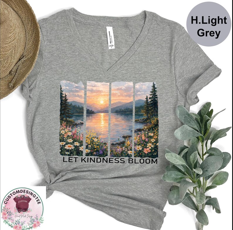 a grey t-shirt with a printed design depicting a serene landscape with a lake, mountains, and a sunset. The text "LET KINDNESS BLOOM" is printed below the image.