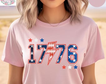 May include: Pink t-shirt with the number "1776" in a patriotic design. The numbers are outlined in blue with white stars, and the lightning bolt is red and white. Red and white stars are above and below the numbers.