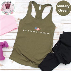 a military green tank top with the text "250 YEARS OF FREEDOM" printed on it, along with an American flag design. The tank top is placed on a wooden surface, and there are some other items, such as a pink headband and a pair of black leggings, visible in the image.