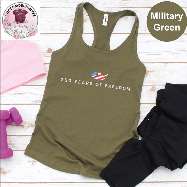 a military green tank top with the text "250 YEARS OF FREEDOM" printed on it, along with an American flag design. The tank top is placed on a wooden surface, and there are some other items, such as a pink headband and a pair of black leggings, visible in the image.
