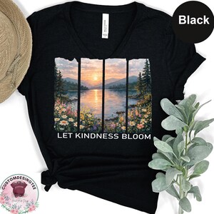 A black t-shirt with a printed design depicting a serene landscape with a lake, mountains, and colorful flowers. The text on the t-shirt reads "LET KINDNESS BLOOM".
