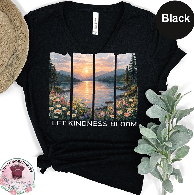 A black t-shirt with a printed design depicting a serene landscape with a lake, mountains, and colorful flowers. The text on the t-shirt reads "LET KINDNESS BLOOM".