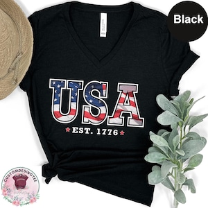 May include: Black V-neck t-shirt with the word "USA" in red, white, and blue, with the American flag design. The text "EST. 1776" is below the word "USA".