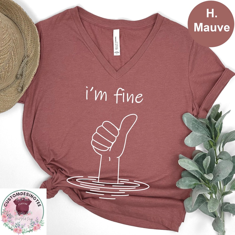 a red v-neck t-shirt with a hand gesture and the text "i'm fine" printed on it. The t-shirt is placed on a white surface, with a straw hat and some greenery visible in the background.