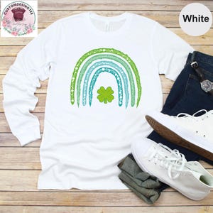 May include: White long-sleeve t-shirt with a green shamrock in a rainbow design. The rainbow is made of green, blue, and white stripes with white stars.