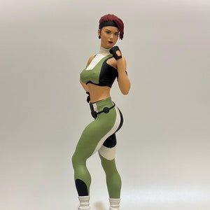 May include: A green and white action figure of a female character with red hair, wearing a green and black top and trousers. The figure is standing on a circular base with a skull and bones underneath.